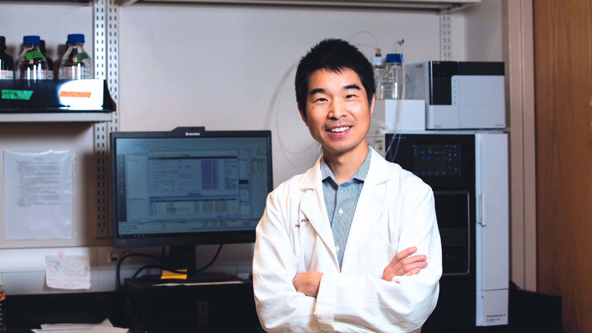 Qingguo Xu smiles in a lab coat with his arms folded in front of lab equipment in his lab.