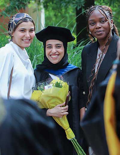 a graduate in academic regalia smiles and holds a bouquet while posing with two friends at an outdoor ceremony