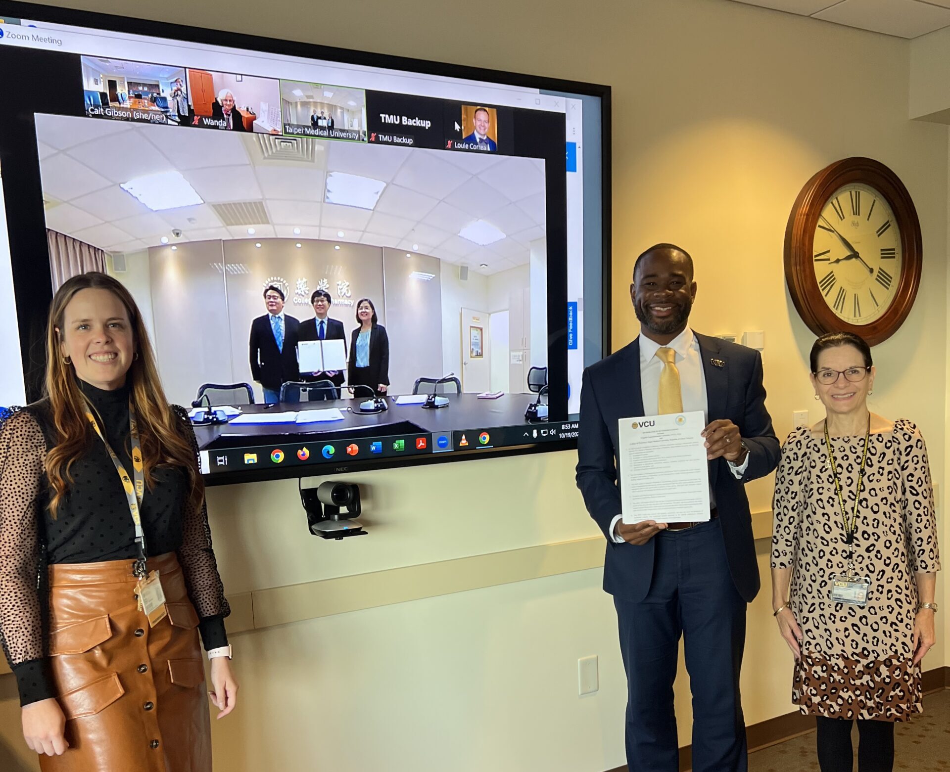 Three individuals, including one holding a memorandum of understanding, pose for a photo while standing next to a projector screen where three more individuals from Taipei Medical University are posing for a photo via video call.